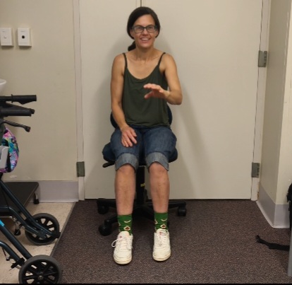 I am sitting on an office chair in a hospital room in front of a white wall and door. My left hand is raised chest height as I perform the hand movements of the assessment as part of the levodopa challenge. My right hand rests on my leg. I am smiling but I am really not happy inside because it is hard.