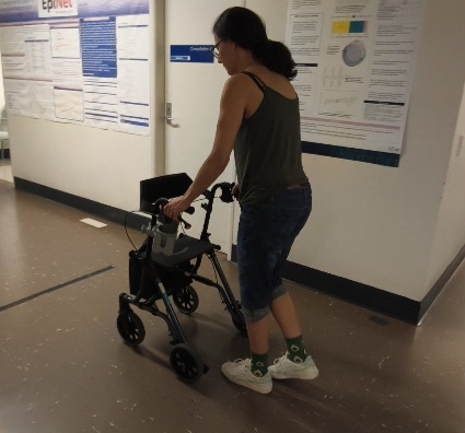 Me holding onto the handles of my walker (mobility device with four wheels and a seat). I am walking down a hospital corridor, bent over slightly and taking small steps.